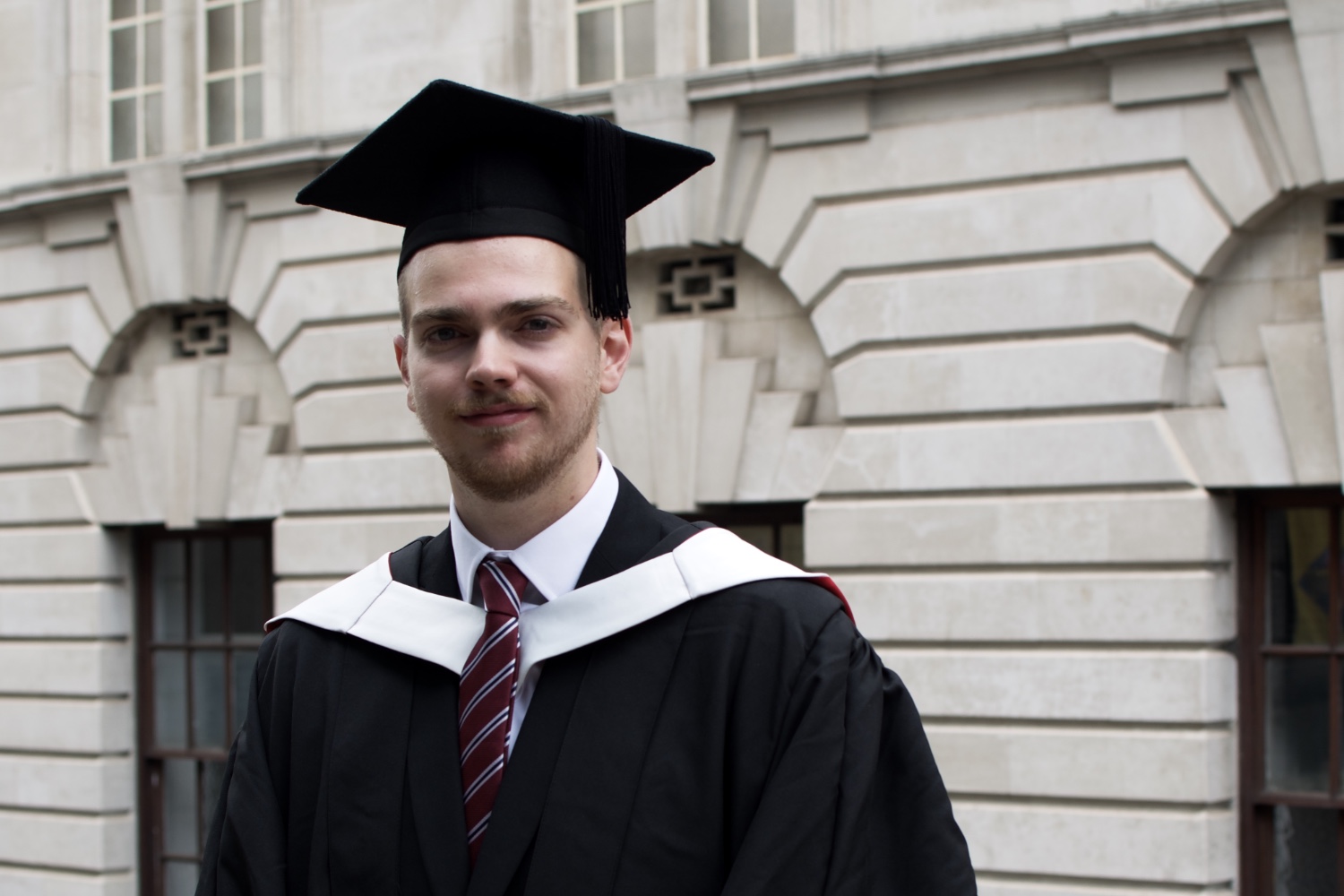 Photograph of Marley Sudbury wearing academic dress at his graduation. He is stood in front of a stone university buidling.